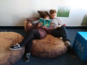 Ein jugendliches Paar sitzt auf einem Sitzkissen und liest, sie hält ein Buch hoch mit dem Titel "It's a Nerd's World"